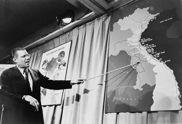 Black and white photo of Defense Secretary Robert McNamara using a pointer to locate where the alleged incident took place. Dated 1965.